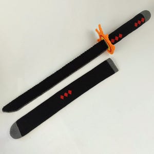 May include: A black crochet sword with a matching sheath. The sword features a black handle with red diamond accents and an orange guard. The sheath also has red diamond accents. The sword and sheath are set against a white background.