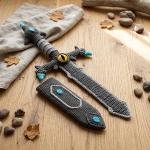 May include: A crocheted toy sword and sheath set. The sword is gray with blue and yellow accents, and the sheath is gray with a blue accent. The set is placed on a wooden surface with leaves and stones.