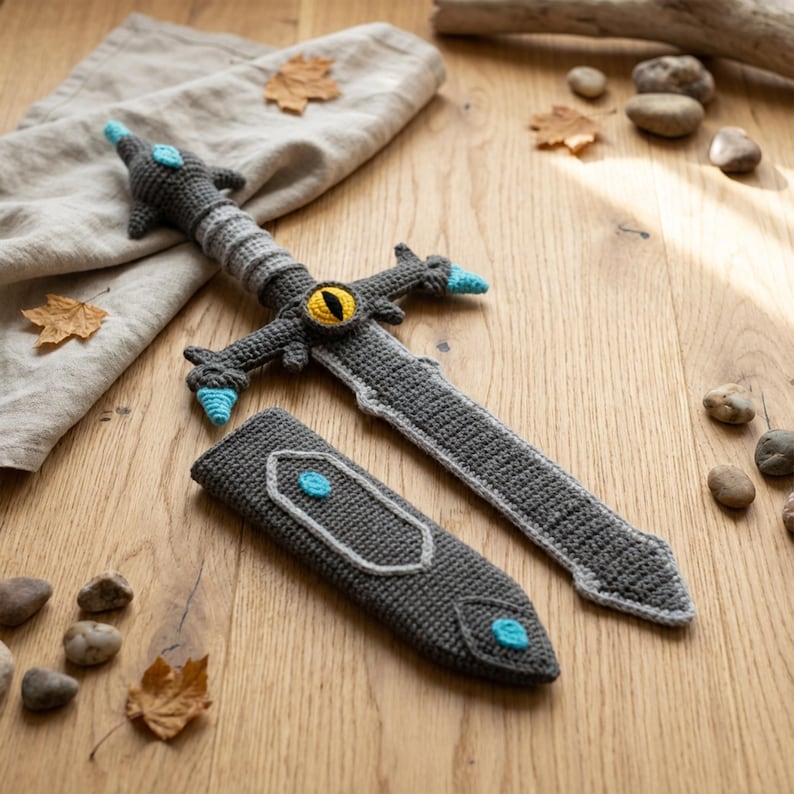 May include: A crocheted toy sword and sheath set. The sword is gray with blue and yellow accents, and the sheath is gray with a blue accent. The set is placed on a wooden surface with leaves and stones.