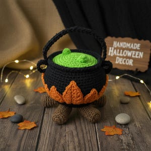 K&ouml;nnte beinhalten: Ein handgeh&auml;kelter Halloween-Kessel mit schwarzem K&ouml;rper, orangefarbenen Akzenten und einem gr&uuml;nen, brodelnden Deckel. Der Kessel hat einen Henkel und steht auf braunen Beinen. Das Holzschild im Hintergrund tr&auml;gt die Aufschrift "HANDMADE HALLOWEEN DECOR".
