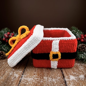 Può includere: Una scatola rossa all'uncinetto con finiture bianche e un design a cintura nera, che ricorda l'abito di Babbo Natale. Il coperchio è aperto, rivelando l'interno. Un fiocco giallo adorna il coperchio. La scatola è su una superficie di legno con neve finta.