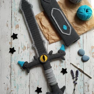 May include: A handmade crochet sword with a matching sheath. The sword is primarily gray with light blue and yellow accents. The handle is wrapped in gray yarn. The sheath is gray with light blue details. The sword is lying on a wooden surface.