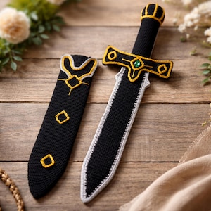 May include: A black and white crochet sword and sheath set with gold detailing. The sword has a green gem and gold features on the hilt and crossguard. The sheath has gold trim and diamond-shaped accents. A decorative item.