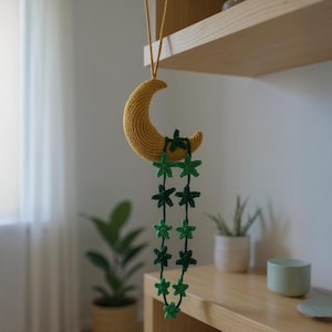 Könnte beinhalten: Gehäkeltes Wandbehang mit einem gelben Halbmond und einer Kette aus grünen sternförmigen Ornamenten. Der Mond wird von einer gelben Schnur gehalten. Die Sterne sind in verschiedenen Grüntönen gehalten.