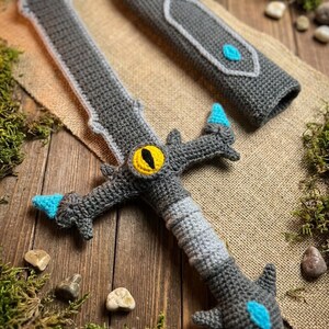 May include: Crocheted gray sword with a matching sheath. The sword features a yellow eye design and blue accents. The handmade toy is displayed on a wooden surface with moss and small stones.