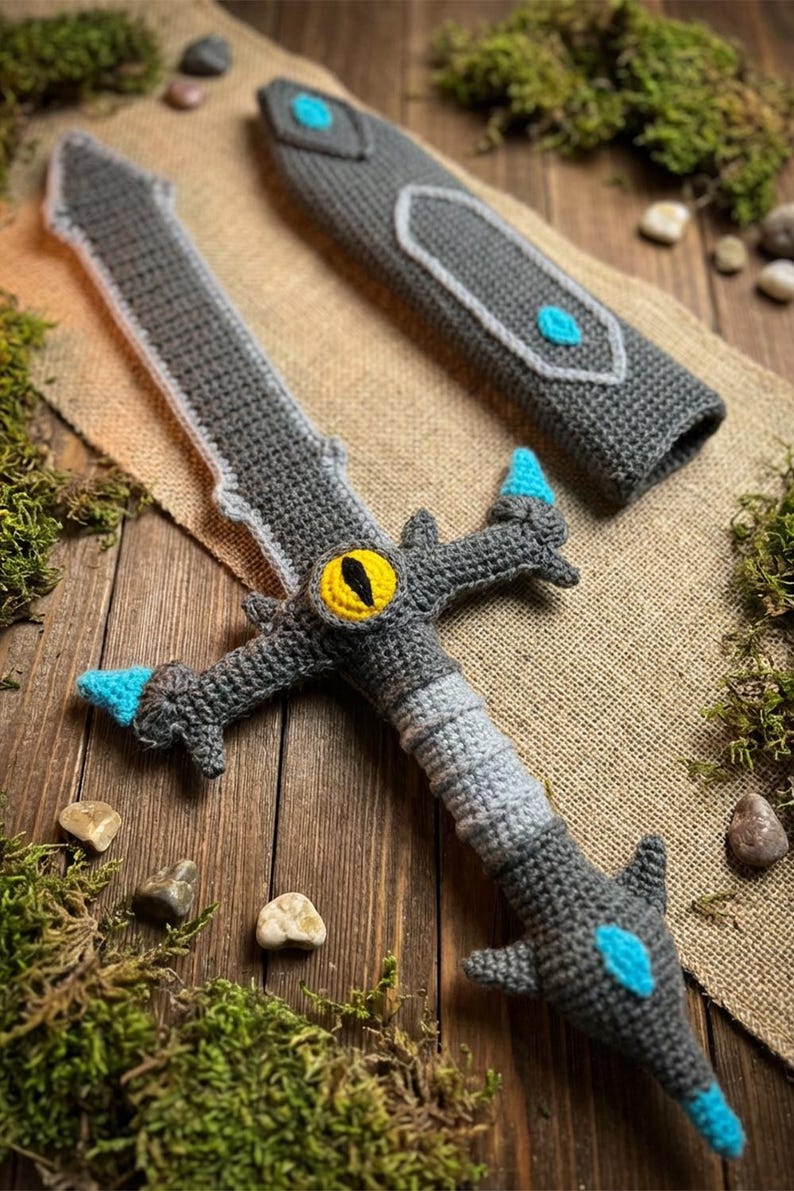 May include: Crocheted gray sword with a matching sheath. The sword features a yellow eye design and blue accents. The handmade toy is displayed on a wooden surface with moss and small stones.