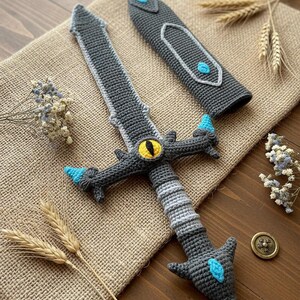 May include: Crocheted sword with a gray blade, a yellow eye, and blue accents. The sword is accompanied by a matching sheath. The handmade fantasy item is displayed on a burlap surface with decorative wheat stalks.