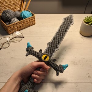 May include: Handmade crochet sword in shades of gray with blue and yellow accents. The sword features a detailed design, including a yellow eye in the center of the crossguard. The handle is held by a person. The sword is a unique, handcrafted item.