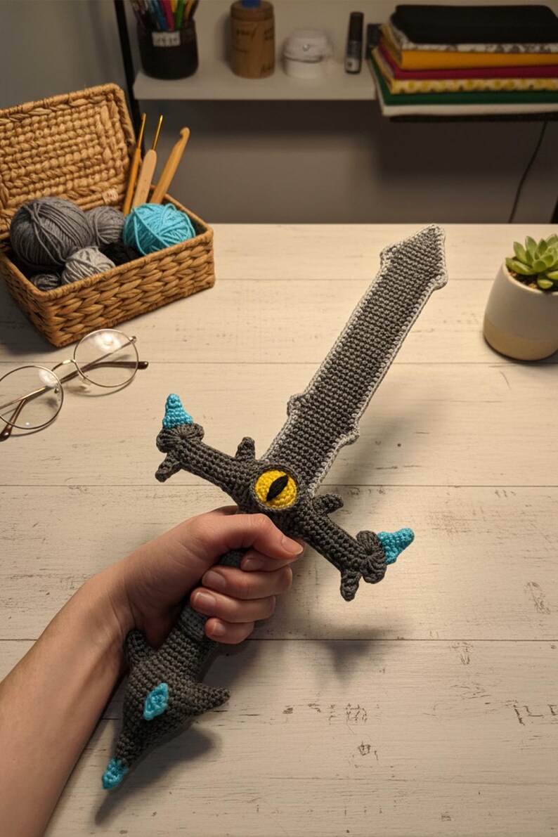 May include: Handmade crochet sword in shades of gray with blue and yellow accents. The sword features a detailed design, including a yellow eye in the center of the crossguard. The handle is held by a person. The sword is a unique, handcrafted item.