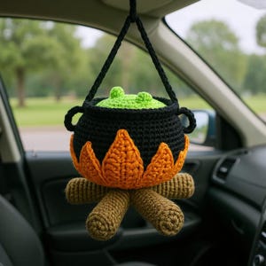 Crocheted hanging amigurumi of a witch's cauldron with bubbling green potion, surrounded by bright orange flames and brown fire logs. The soft plush toy is suspended from a car mirror, serving as a spooky Halloween-themed charm. The black pot features tiny side handles, and the playful design evokes a cozy campfire or witch's brew. Ideal for car decor or handmade gifts. Pattern sold as a digital PDF crochet tutorial.
