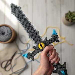 May include: A handmade crochet sword in shades of gray, with yellow and blue accents. The sword features a detailed design, including a yellow eye in the center and blue tips. The craft item is held against a light wooden background.