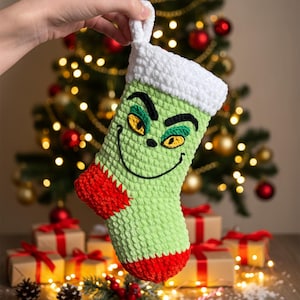 Puede incluir: Un calcetín navideño verde y rojo con un diseño de la cara del Grinch. El calcetín está tejido a ganchillo con un puño blanco y un talón y puntera rojos. La cara del Grinch está bordada con hilo negro, verde y amarillo.