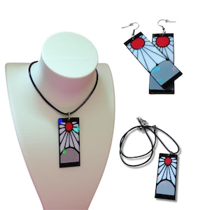 Op de afbeelding: Een ketting- en oorbellen set met een rechthoekige hanger met een rood zonontwerp, witte stralen en een grijze en zwarte rand. De ketting heeft een zwart koord en de oorbellen hebben zilveren haken. De sieraden worden getoond op een witte mannequin.