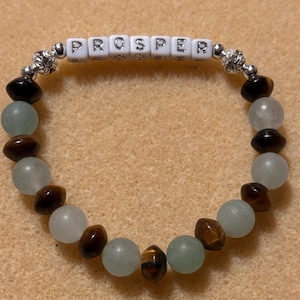 Puede incluir: Una pulsera de cuentas con la palabra "PROSPER" escrita con cuentas de letras blancas. La pulsera presenta una mezcla de cuentas azul claro, marrón y negras, con detalles plateados.