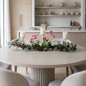 Könnte beinhalten: Ein runder, hellfarbiger Esstisch mit einem dekorativen Mittelstück. Das Mittelstück besteht aus drei weißen Kerzenhaltern mit brennenden Kerzen, rosa und weißen Blumen und grünem Laub. Der Tisch ist von gepolsterten Stühlen umgeben.