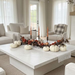 Puede incluir: Un salón decorado para el otoño. Una mesa de centro cuadrada presenta calabazas blancas, hojas de otoño y velas naranjas. La habitación incluye muebles blancos, tonos neutros y cortinas transparentes. La mesa mide aproximadamente 1,5 metros cuadrados.