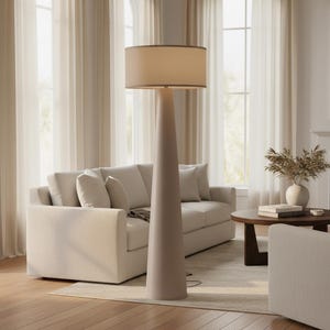 May include: A tall, beige floor lamp with a conical base and a cylindrical lampshade. The lampshade is a lighter beige color with a darker trim. The lamp is in a living room setting with a sofa and a round coffee table.