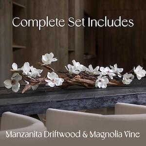 Puede incluir: Un conjunto completo de flores de vid de magnolia blancas dispuestas sobre una rama de madera flotante de manzanita natural. Las flores y la rama se muestran sobre una mesa de madera oscura.