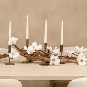 Puede incluir: Un centro de mesa decorativo con cinco velas blancas y delgadas encendidas en candelabros de bronce. Las velas están dispuestas sobre una rama de madera natural adornada con flores de magnolia blancas.