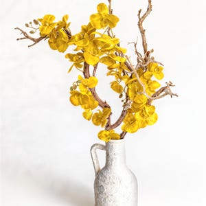 May include: A white, textured vase with a handle holds a display of bright yellow flowers and brown branches. The arrangement is set against a clean white background, creating a striking contrast. The vase is approximately 10 inches tall.