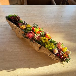 May include: A long, beige, textured planter filled with a variety of colourful artificial succulents. The succulents include shades of green, yellow, red, and pink. The planter is placed on a light-coloured surface.