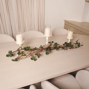 Könnte beinhalten: Ein Holzast mit grünen Blättern und drei weißen Kerzen in Glasbehältern auf einem weißen Tisch mit weißen Stühlen.