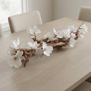 May include: A decorative floral arrangement featuring white magnolia blossoms attached to a natural-looking, brown, wooden branch. The arrangement is placed on a light-coloured wooden table, suitable for home decor.