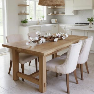 Może przedstawiać: Jasny drewniany stół jadalny z czterema tapicerowanymi krzesłami. Stół jest prostokątny i ma naturalne wykończenie drewna. Na stole leży dekoracyjna gałązka z białymi kwiatami. Krzesła mają jasną tapicerkę i drewniane nogi.