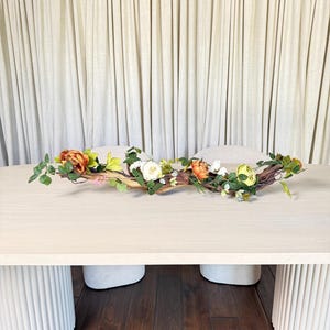 May include: A decorative floral arrangement featuring a natural branch adorned with faux flowers in shades of orange, white, and yellow, with green foliage. The arrangement is placed on a light-colored wooden table. The table has white, cylindrical legs.