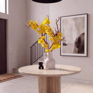 May include: A round, light-colored stone table with a white textured vase holding yellow flowers and branches. A small black decorative object sits on the table. A framed abstract art piece hangs on the wall.