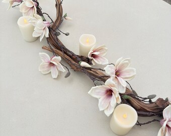 May include: A white table centerpiece with a driftwood branch adorned with white magnolia blossoms and three white pillar candles.