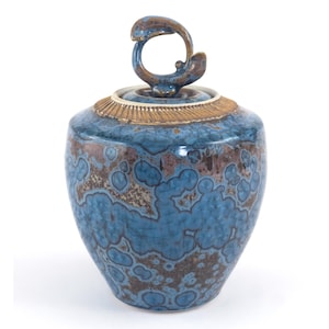 Puede incluir: Un jarrón de cerámica azul con tapa y asa. El jarrón tiene una superficie texturizada con detalles en marrón y blanco. La tapa está rematada con un asa en forma de lazo de color marrón oscuro. El jarrón tiene una banda dorada alrededor de la parte superior.