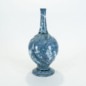 Puede incluir: Un jarrón decorativo de cerámica azul y blanco con un cuerpo bulboso y un cuello delgado. El jarrón presenta un patrón arremolinado y se asienta sobre una base de pedestal. La altura del jarrón es de unos 30 cm.