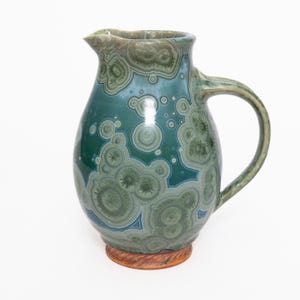 Puede incluir: Jarra de cerámica verde y azul turquesa con un patrón único de círculos concéntricos y formas orgánicas. Tiene un pico y un asa curva. Se apoya sobre una base circular marrón. Altura: aproximadamente 25 cm.