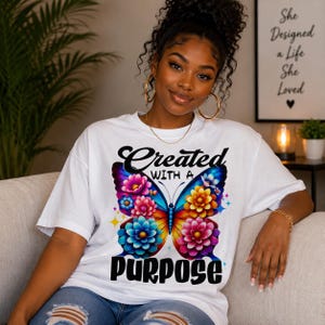 Puede incluir: Camiseta blanca con un diseño de mariposa colorida y la frase "Created with a Purpose". La mariposa presenta patrones florales azules, rosas, amarillos y naranjas. La persona lleva pendientes de aro dorados.