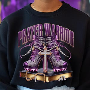 May include: A black sweatshirt with the words "PRAYER WARRIOR" in gold lettering. The design features a pair of purple high-heeled shoes with rhinestones, a gold cross, and a gold chain on a bible.