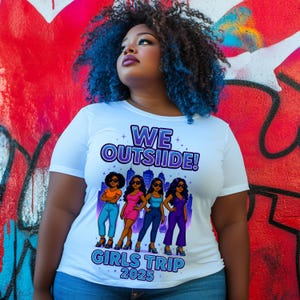 May include: White t-shirt with a graphic of four women in front of a city skyline. The text "WE OUTSIDE! GIRLS TRIP 2025" is printed on the shirt.