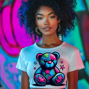 Pode incluir: Uma camiseta branca de corte curto com um design de urso de pelúcia de patchwork colorido. O urso de pelúcia tem estrelas rosa nas patas e um nariz azul. A camiseta tem gola redonda e mangas curtas.