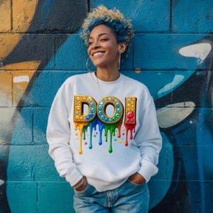 May include: White sweatshirt with the word "DOI" in a jeweled, dripping design. The letters are outlined in gold and filled with colorful gems. Rainbow-colored drips and stars cascade down the front of the sweatshirt.