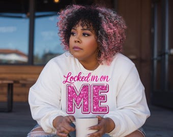 May include: A white sweatshirt with the text "Locked in on ME" in pink glitter lettering. The sweatshirt is paired with ripped blue jeans. A white mug is held in the hands.