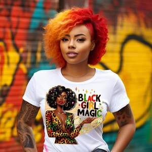 Op de afbeelding: Wit T-shirt met de tekst "BLACK GIRL MAGIC" en een afbeelding van een zwarte vrouw in een jurk met luipaardprint. De vrouw heeft krullend zwart haar en gouden oorringen. De achtergrond is een kleurrijke graffiti-muur.