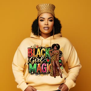Op de afbeelding: Een lichtgele hoodie met de tekst "BLACK Girl MAGIC" in een kleurrijk ontwerp. De hoodie heeft een afbeelding van een vrouw in een luipaardprint jurk. De vrouw draagt een gouden kroon en oorringen.