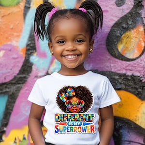 以下が含まれることがあります： 白いTシャツを着た少女。「DIFFERENT IS MY SUPERPOWER」の文字入り。Tシャツには、巻き毛、ハート型のメガネ、カラフルなリボンをした女の子のグラフィックが描かれています。背景はカラフルな落書きの壁です。