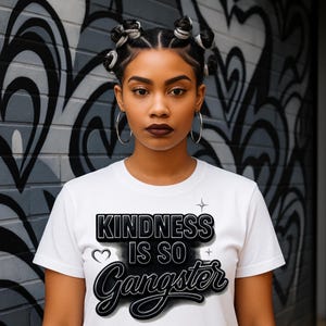 May include: A white t-shirt with the phrase "KINDNESS IS SO Gangster" in a black, outlined font. The model is wearing large silver hoop earrings. The background is a gray and white graffiti wall.