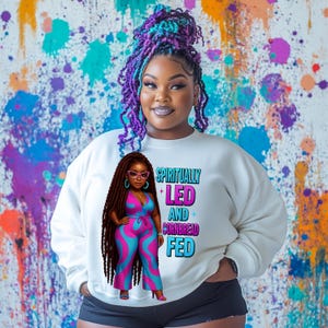 Puede incluir: Sudadera blanca con una ilustración de dibujos animados de una mujer con rastas largas que lleva un mono rosa y azul. El texto "Spiritually Led and Cornbread Fed" está impreso en la sudadera.