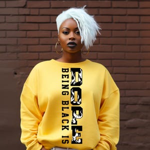 Puede incluir: Una sudadera amarilla con el texto "Being Black Is Dope" en un diseño vertical. La sudadera tiene mangas largas y cuello redondo. El texto es en blanco y negro.