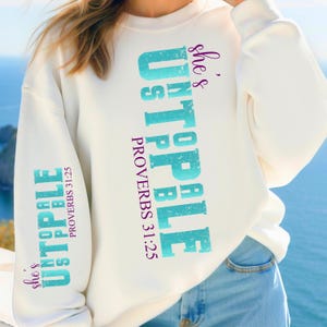 May include: White sweatshirt with the text "She's unstoppable" in purple and blue lettering. The text "Proverbs 31:25" is also printed in purple.