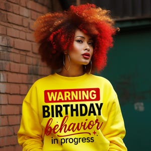 May include: A woman with bright red and orange curly hair and large hoop earrings is wearing a yellow sweatshirt. The sweatshirt has the words "WARNING BIRTHDAY behaviour in progress" printed on it in black and red.