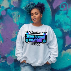 May include: A light blue sweatshirt with the text "Caution Zero Sugar Coating Period" in a black rectangle with blue and purple accents. The sweatshirt is worn by a person with blue and purple hair.