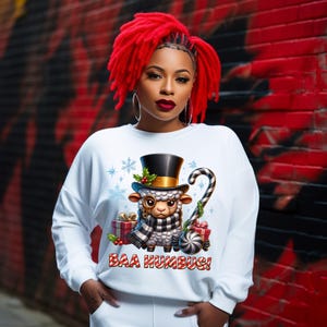 May include: White sweatshirt with a graphic of a sheep wearing a top hat, scarf, and glasses, holding a candy cane. The text "BAA HUMBUG!" is below the sheep. The background is a red and black brick wall.